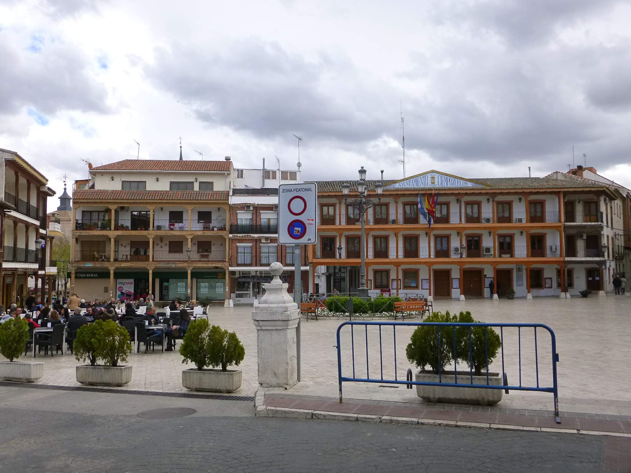 Ein belebter Stadtplatz mit Menschen auf Stühlen sitzend und Stehenden, umgeben von Topfpflanzen, Metallabsperrungen, Straßenlaternen mit Fahnen und einem Schild, Gebäuden mit Fenstern und einem bewölkten Himmel.