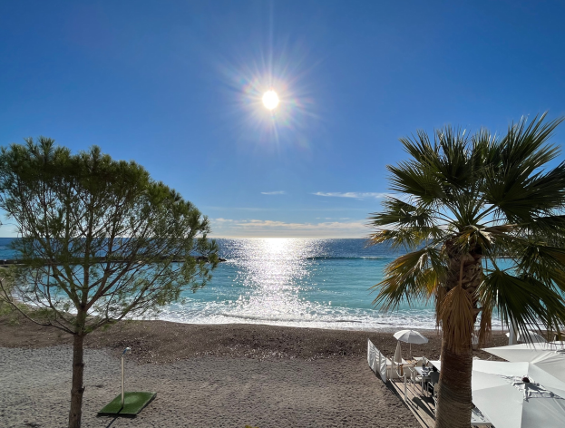 Ein sonniger Strand mit Palmen, Schirmen und üppiger Vegetation an der französischen Riviera unter einem strahlend blauen Himmel.