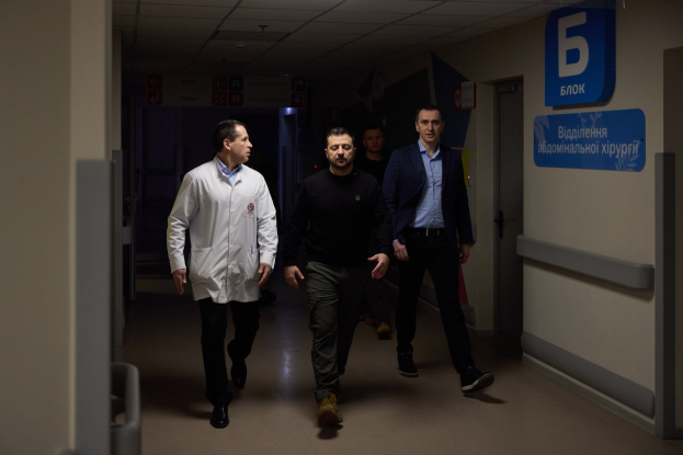 Eine Gruppe von Männern geht einen Krankenhausflur entlang, mit beleuchteter Decke, einer Tür auf der rechten Seite und Tafeln mit Text auf der linken Seite, die besorgt aussehen.