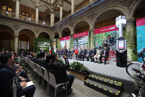 Eine Gruppe von Menschen, die in einem Gebäude auf Stühlen sitzen, mit einem Mann auf der Bühne, der ein Mikrofon hält, Fahrräder auf der rechten Seite geparkt und verschiedene Elemente wie Plakate, Pflanzen, Säulen, Geländer, Lampen, Fenster und eine Wand im Hintergrund, was eine Pressekonferenz für das CDMX-Nachhaltigkeitsenergiegipfel vermuten lässt.