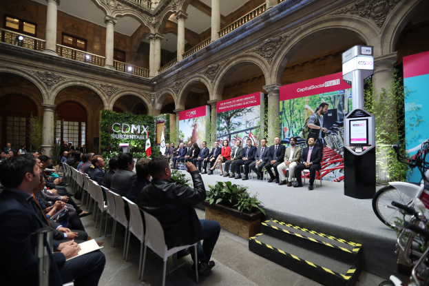Eine Gruppe von Menschen, die in einem Gebäude auf Stühlen sitzen, mit einem Mann auf der Bühne, der ein Mikrofon hält, Fahrräder auf der rechten Seite geparkt und verschiedene Elemente wie Plakate, Pflanzen, Säulen, Geländer, Lampen, Fenster und eine Wand im Hintergrund, was eine Pressekonferenz für das CDMX-Nachhaltigkeitsenergiegipfel vermuten lässt.