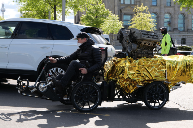 Ein Mann im Rollstuhl mit einem großen Motor auf dem Rücken, umgeben von Fahrzeugen auf einer Straße mit Bäumen, Gebäuden, Masten und einem klaren blauen Himmel im Hintergrund, der eine schwarze Jacke und eine Mütze trägt und ein Objekt hält.