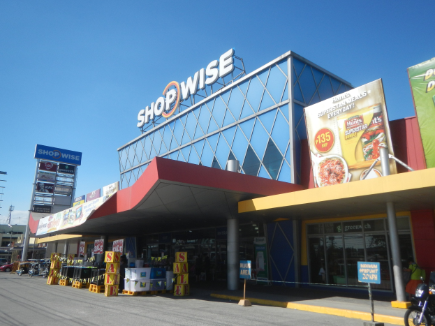Verkaufsfront eines Shopwise-Supermarkts mit einem vor der Tür geparkten Motorrad, umgeben von Fahrzeugen, Pfählen, Plakaten und anderen städtischen Objekten, mit Gebäuden, Bäumen und einem klaren blauen Himmel im Hintergrund.