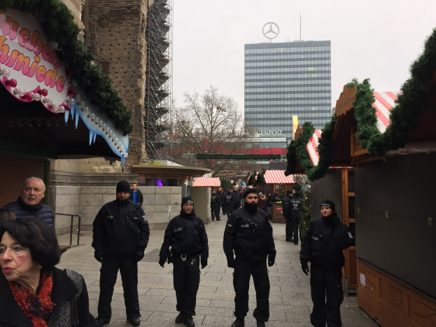 Polizisten in schwarzen Uniformen stehen vor einem geschmückten Weihnachtsmarkt in Berlin, mit festlichen Dekorationen, Gebäuden, Bäumen und einem klaren blauen Himmel im Hintergrund.