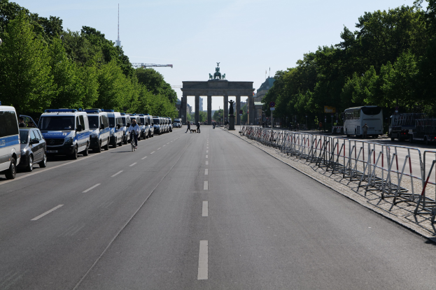 Lange Reihe von Polizeiwagen, die auf der Seite einer Straße vor dem Brandenburger Tor in Berlin, Deutschland, geparkt sind, mit Menschen auf Fahrrädern und auf der Straße stehend, Barrieren, Bäumen und einem Bogen mit Statuen im Hintergrund.