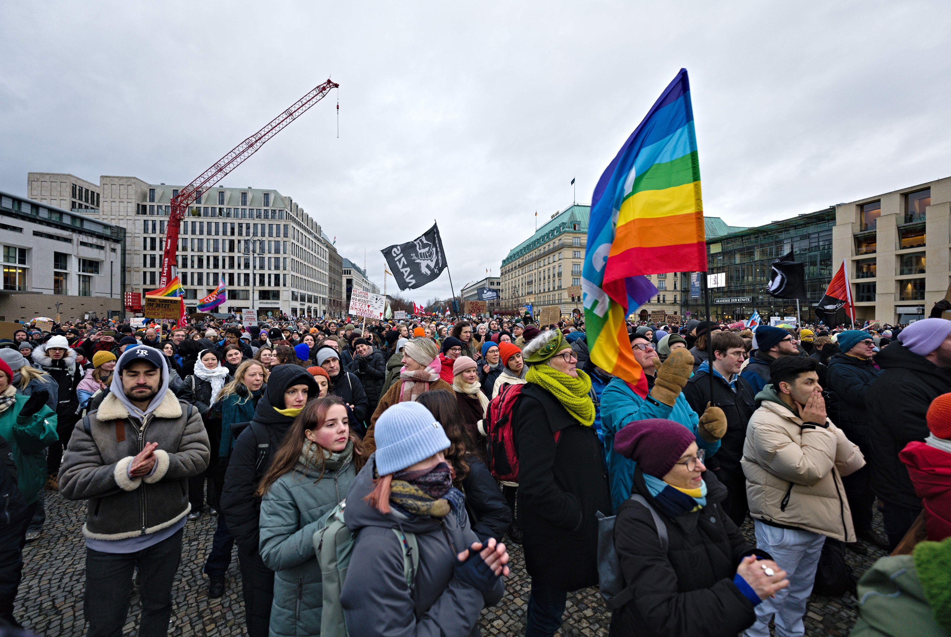 Große Gruppe von Menschen bei einer LGBTQ+-Rechtsdemo in Berlin, die Fahnen und Schilder halten, mit Gebäuden, einem Kran und bewölktem Himmel im Hintergrund.