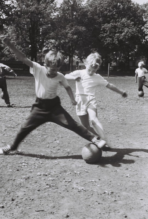 Schwarzes und weißes Foto von jungen Jungs, die Füüball auf einem Feld mit Bäumen und einem klaren Himmel im Hintergrund spielen.