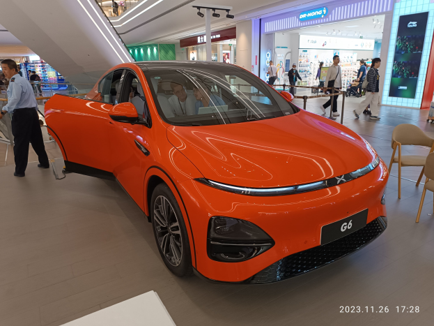 Orangefarbener Elektroauto in einer Ausstellungshalle umgeben von Menschen, Stühlen, einem Wagen und Geschäften im Hintergrund.