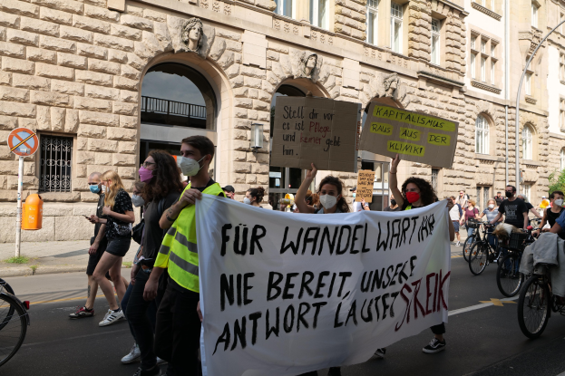 Eine Gruppe von Menschen marschiert bei einer Demonstration auf einer Stadtstraße, einige halten Schilder und andere fahren Fahrräder, mit einem historischen Gebäude im Hintergrund, das Rundbogenfenster, Säulen und Skulpturen aufweist.