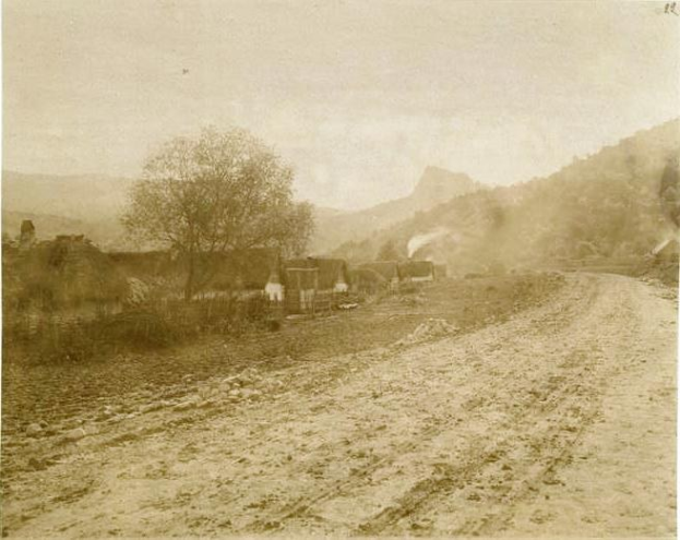 Ein altes Schwarz-Weiß-Foto einer Schotterstraße, die sich durch eine ländliche Landschaft schlängelt, mit Häusern, Bäumen und Bergen im Hintergrund und einem klaren Himmel.