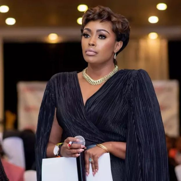 Eine Frau in einem schwarzen Kleid mit Mikrofon und Papier, die mit einem sitzenden Publikum diskutiert.