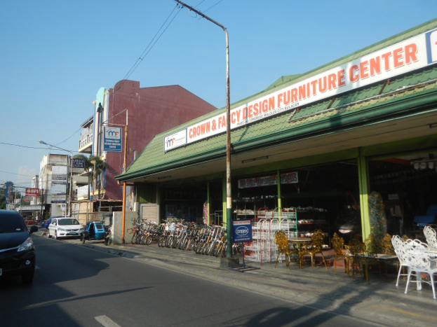 Eine belebte Stadtstraße mit fahrenden Fahrzeugen, parkenden Fahrrädern, Gebäuden mit Schildern, Laternenmasten und Stromleitungen, mit einem Möbelgeschäft im Vordergrund und Himmel im Hintergrund.