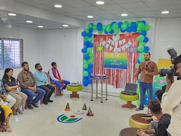 Eine Gruppe von Menschen, die auf Stühlen sitzen und einer Menge gegenüber sind, mit einem Redner, der ein Mikrofon hält, einem Laptop auf einem Hocker, einem Tisch mit Gegenständen, einem Banner, Luftballons, Vorhängen, einem Fenster und Deckenleuchten, was eine 9. Geburtstagsfeier vermuten lässt.