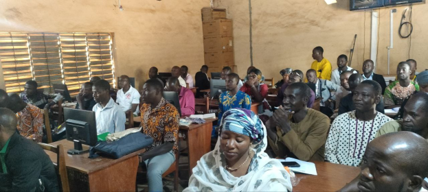 Eine Gruppe von Menschen sitzt an Tischen in einem Klassenzimmer mit Monitoren, Taschen, Büchern und Papieren und nimmt an einer Techniktrainingssitzung teil; ein Fenster links und Kartons mit einem Fernseher an der Wand im Hintergrund.