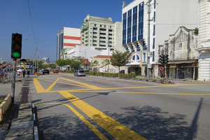 Stadtstraßenszene mit Fahrzeugen, Fußgängern, Ampeln, Strommasten, Grünflächen und Gebäuden unter einem klaren blauen Himmel.