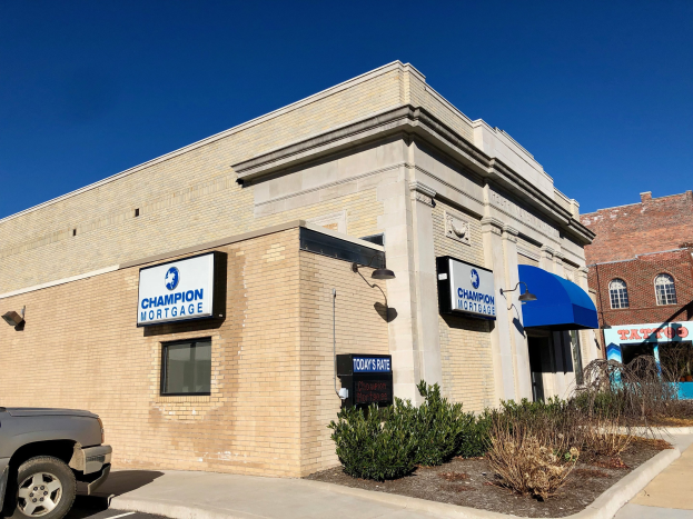 Ein zweistöckiges Gebäude mit vernagelten Fenstern, einem geparkten Auto, Topfpflanzen und Bäumen unter einem bewölkten Himmel, das ein "Champion Mortgage, Inc." Schild zeigt.