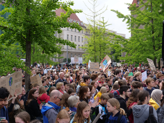 Eine große Menschenmenge protestiert vor einem Gebäude in Berlin und hält Schilder hoch, mit Bäumen, Fahrzeugen und einem Lautsprecher im Hintergrund.