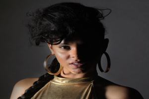 Eine Frau in einem goldenen Kleid und großen Creolen steht selbstbewusst vor einer Wand, ihr glatter Bob-Haarschnitt und das natürliche Make-up unterstreichen ihren entschlossenen Gesichtsausdruck, während sie direkt in die Kamera schaut.