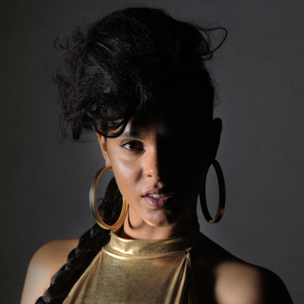 Eine Frau in einem goldenen Kleid und großen Creolen steht selbstbewusst vor einer Wand, ihr glatter Bob-Haarschnitt und das natürliche Make-up unterstreichen ihren entschlossenen Gesichtsausdruck, während sie direkt in die Kamera schaut.