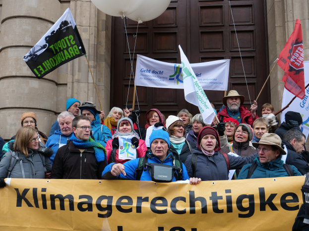 Eine Gruppe von Menschen steht vor einem Gebäude mit Säulen und einer Tür, hält ein Banner mit Text, Fahnen mit Stangen und Ballons und nimmt an einem Klimastreik in Deutschland teil.