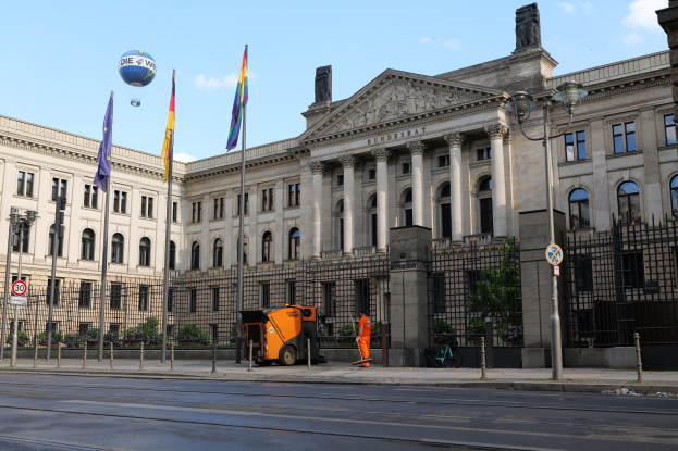 Großes modernes Regierungsgebäude mit vielen Fenstern, umgeben von Straßeninfrastruktur, Füügängern und Bäumen, mit einem bewölkten Himmel im Hintergrund.