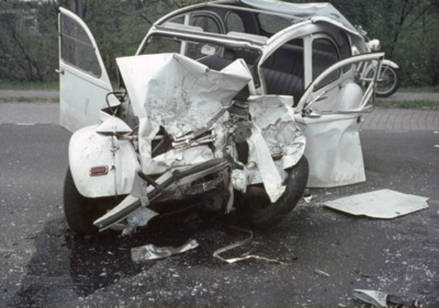 Schwer beschädigte Frontpartie eines Autos am Straßenrand mit Bäumen und Gebäude im Hintergrund, weiteres Fahrzeug in der Ferne sichtbar.