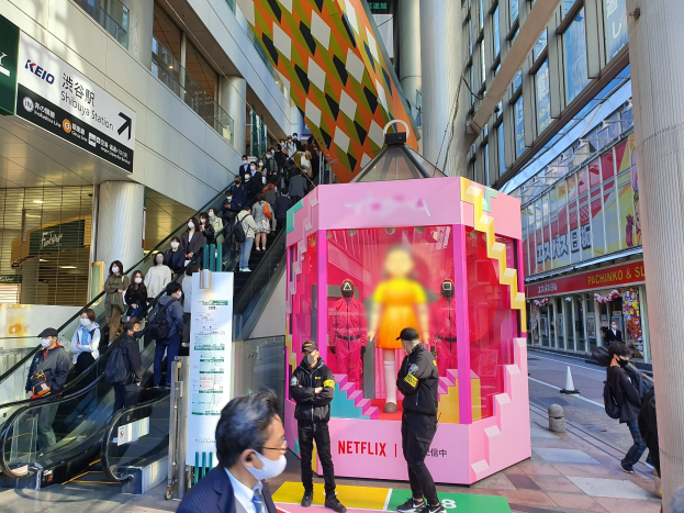 Gruppe von Menschen vor einem Gebäude mit Glaswänden und Texttafeln, einige tragen Masken, mit einer Rolltreppe im Hintergrund und einer pinken Box mit der Aufschrift "Netflix's new Netflix Booth in Tokyo" mit Schaufensterpuppen.