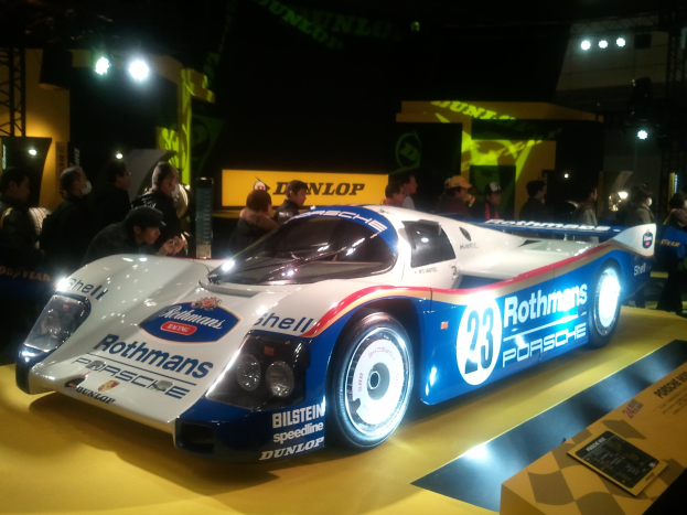 Blauer und weißer Porsche 917K Rennauto auf der 24-Stunden-Rennen von Le Mans, umgeben von Menschen in farbenfrohen Kleidern mit Schildern und Lichtern im Hintergrund.