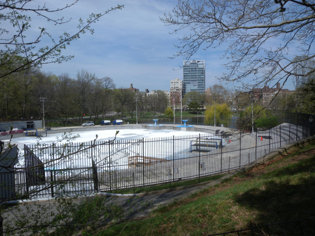 Eisstadion in einem Park umgeben von einem Metallzaun, Gras, Bäumen, Fahrzeugen, Straßenlaternen, einem Gewässer, Gebäuden und einer bewölkten Himmel.