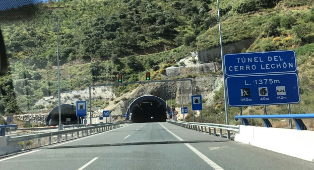 Ein Auto fährt durch einen Tunnel neben einer Straße mit Geländern, Schildern, Pfählen, Verkehrszeichen, Bäumen, Felsen und einem klaren blauen Himmel.