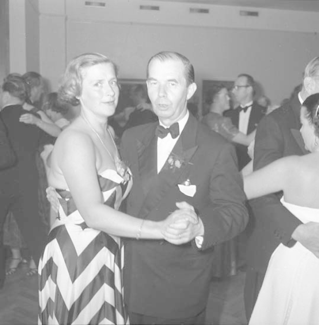 Ein Mann in einem schwarzen Blazer, weißem Hemd und schwarzen Hosen tanzt mit einer Frau in einem unbekannten Outfit bei einem formellen Ereignis, indem sie sich an den Händen halten und zur Musik schwenken, vor einer Wand als Hintergrund.