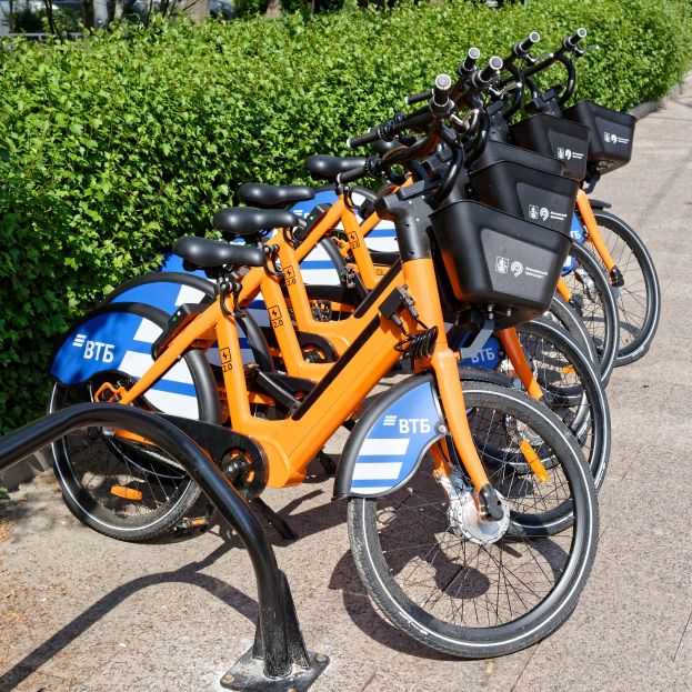 Eine Reihe von orange und blauen Fahrrädern, die nebeneinander auf der Straße geparkt sind, mit Pflanzen, Bäumen und einem Gebäude im Hintergrund.