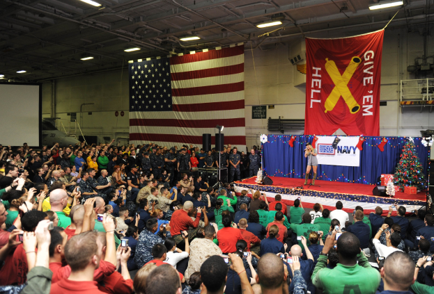 Große Menschenmenge vor einer Bühne mit einer Frau mit Mikrofon, einem geschmückten Weihnachtsbaum, einem Banner, Lautsprechern, einem Bildschirm, einer Flagge, einer Wand, Deckenlampen und Metallstangen, wahrscheinlich während einer US-Navy-Weihnachtsfeier.