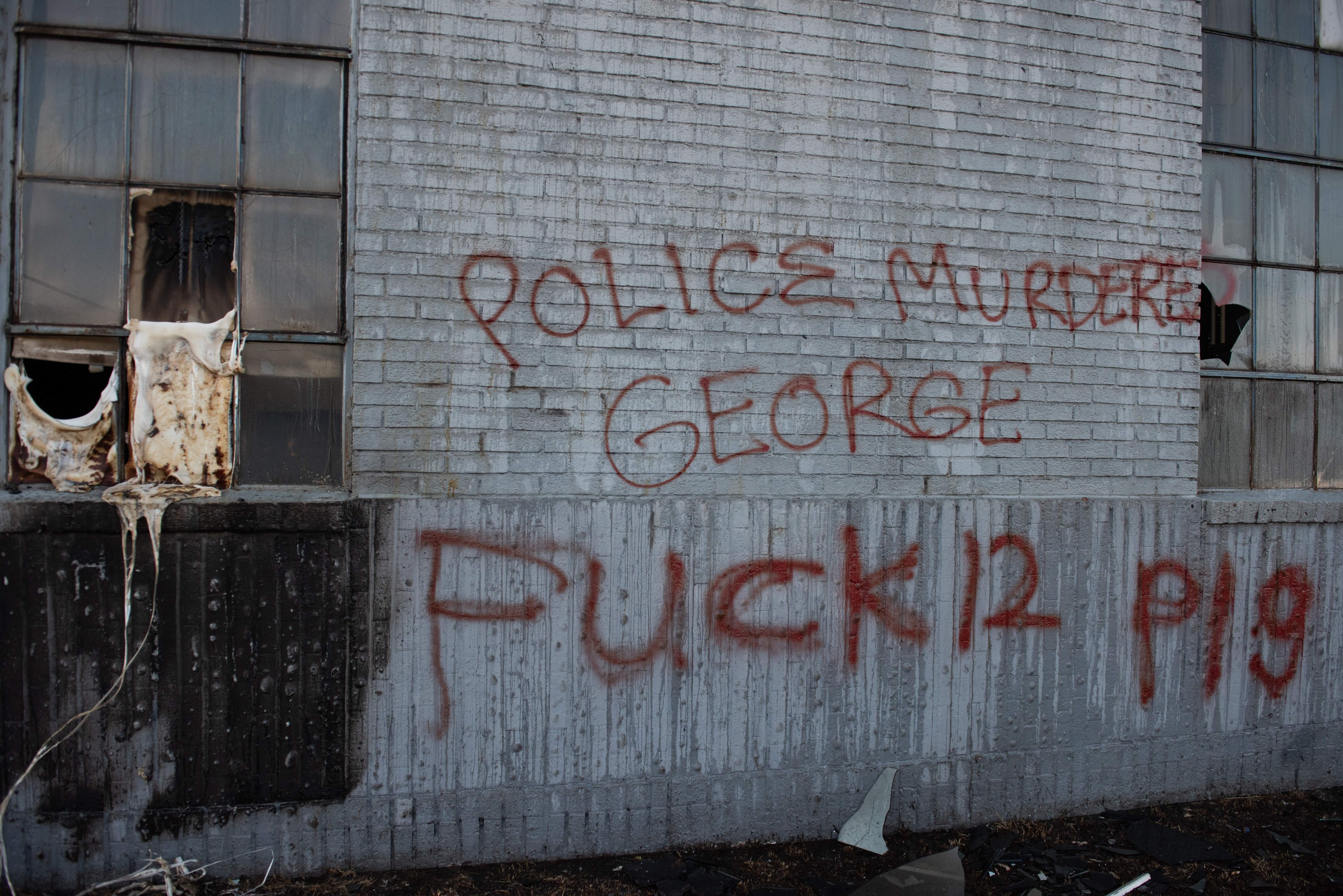 Graffiti an einer Gebäudewand mit der Aufschrift "Polizeimord George Fuck 12 P19" und Fenstern sowie verstreuten Gegenständen davor.