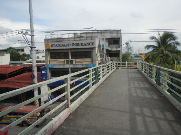 Eine Brücke mit Geländern, Fahrzeugen, Gebäuden, Bäumen, Strommasten mit Kabeln und einem bewölktem Himmel im Hintergrund, mit einem Gebäude an der Seite mit einer "Standard Insurance"-Aufschrift.