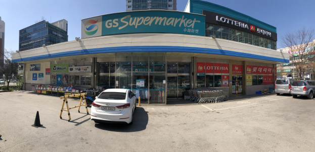 Ein Auto vor einem "GS Supermarkt"-Laden mit anderen Fahrzeugen, Verkehrskegeln und verstreuten Gegenständen auf der Straße, vor dem Hintergrund von Gebäuden, Bäumen und einem klaren blauen Himmel.