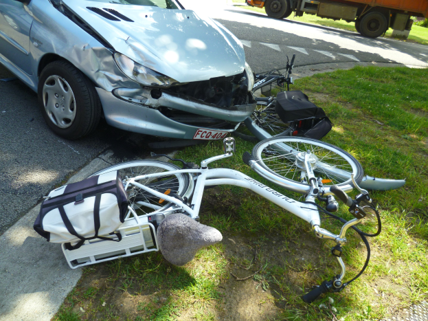 Auto am Straßenrand mit einem Fahrrad in der Nähe auf dem Gras liegend, Bäume und ein klarer blauer Himmel im Hintergrund.