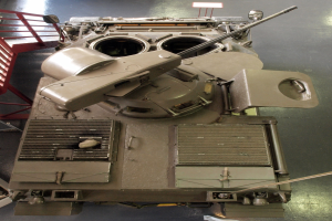 Ein großer Militärpanzer mit sichtbaren Ketten und Turm, der auf einem Boden mit einem Metallgeländer auf der linken Seite positioniert ist.