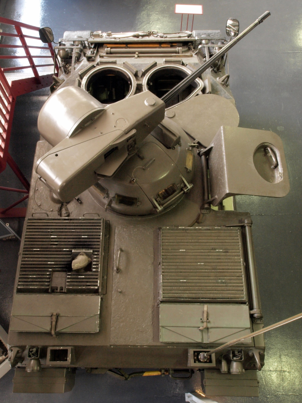 Ein großer Militärpanzer mit sichtbaren Ketten und Turm, der auf einem Boden mit einem Metallgeländer auf der linken Seite positioniert ist.