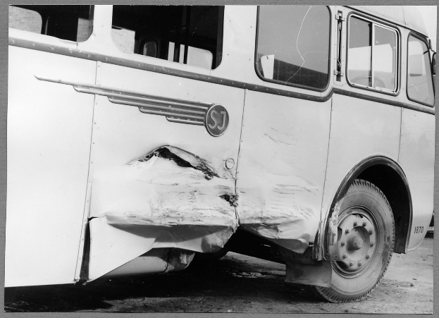 Schwarzes und weißes Foto eines verunglückten Busses mit einer eingedrückten Front, zersplitterten Fenstern und einer offenen Fahrertür, die das Innere zeigt.