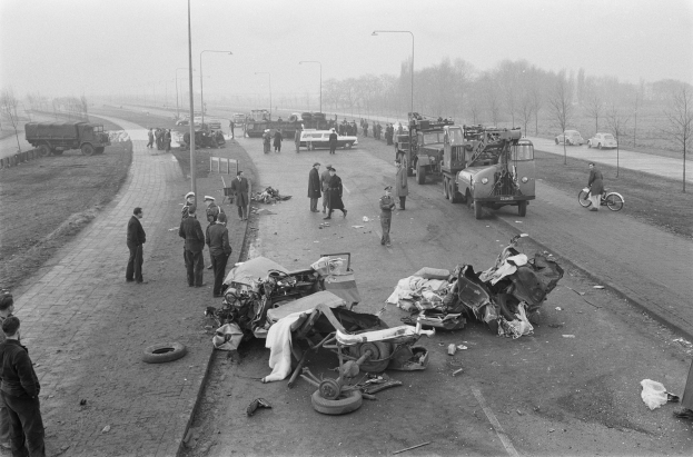 Schwarze und Weiße Szenerie eines Autounfalls am Rande einer Straße mit mehreren Fahrzeugen und einer Gruppe von Menschen in der Nähe, Lichtmäste, Bäume und Himmel im Hintergrund.