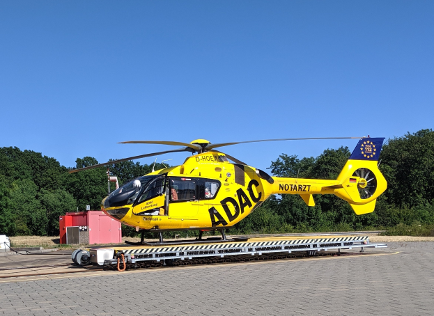 Gelbes Hubschrauber auf einem Flughafenvorfeld mit Bäumen und einem klaren blauen Himmel, umgeben von Gegenständen, die wahrscheinlich mit Luftrettungsdiensten in Deutschland zu tun haben.