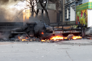 Ein Lkw brennt am Straßenrand, mit Rauch, der daraus aufsteigt, während Bäume, Gebäude und ein klarer Himmel im Hintergrund zu sehen sind.