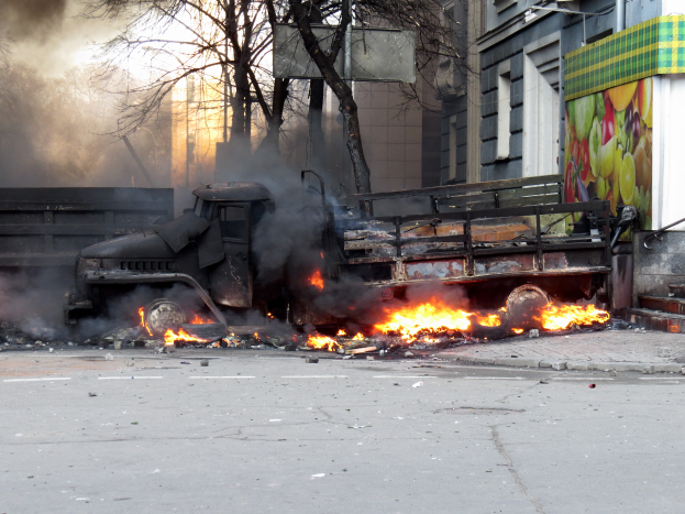 Ein Lkw brennt am Straßenrand, mit Rauch, der daraus aufsteigt, während Bäume, Gebäude und ein klarer Himmel im Hintergrund zu sehen sind.