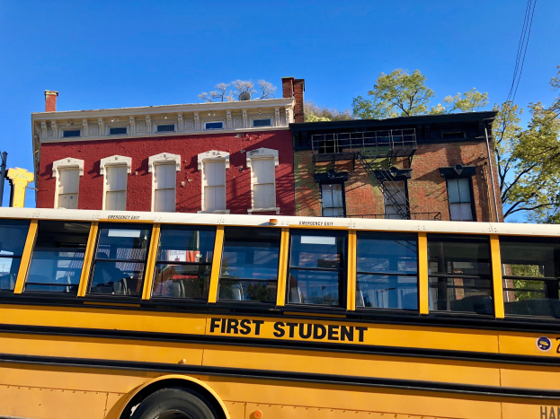 Gelber Schulbus mit der Aufschrift "First Student" vor einem roten Backsteingebäude geparkt, mit ein paar Menschen drinnen, Bäumen und einem klaren blauen Himmel im Hintergrund.