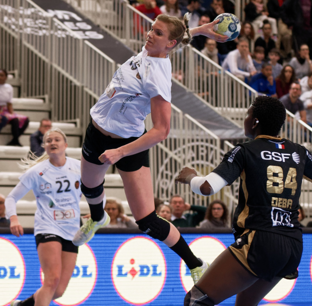 Frauen spielen Handball auf einem Platz mit einem Ball in der Luft, Zuschauer im Hintergrund und einem "Handball-Weltmeisterschaft 2019 - Tag 2"-Schild.