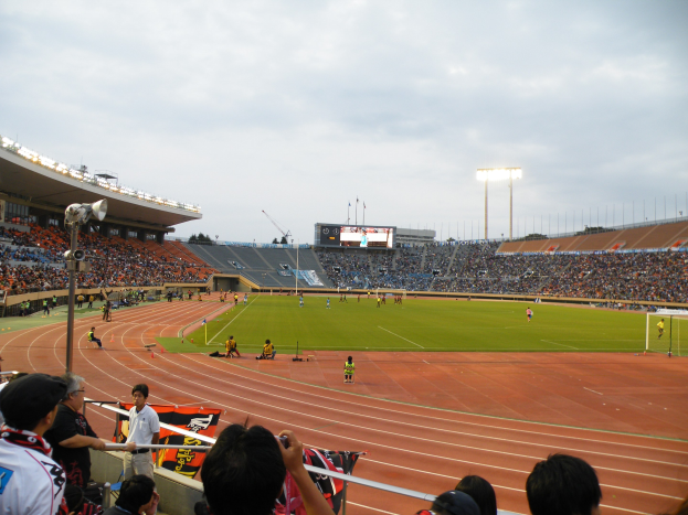 Großes Stadion mit Zuschauern bei einem Fußballspiel, einige sitzen und andere stehen oder halten Kameras, grasbewachsener Platz mit einem Tor, Flutlichtmasten, ein Bildschirm und eine bewölkte Himmelslandschaft im Hintergrund.