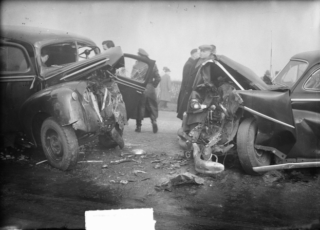 Schwarz-weiß-Foto von zwei beschädigten Autos am Straßenrand mit Menschen in der Nähe und einer Wand im Hintergrund.