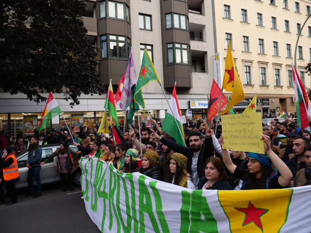 Eine große Gruppe von Menschen marschiert mit Flaggen und Transparenten durch eine Straße, mit einem geparkten Auto auf der rechten Seite und einem Baum auf der linken Seite, vor einem Hintergrund von Gebäuden mit Fenstern und Namensschildern, was auf eine algerische Demonstration hinweist.