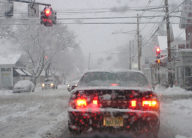 Eine schneebedeckte Straße mit wenigen Fahrzeugen, gesäumt von Häusern und Bäumen auf beiden Seiten.